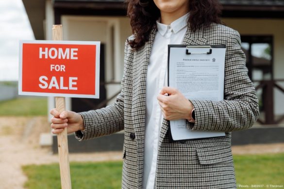 Frau steht vor einem Haus und hält ein rotes Schild mit der Aufschrift "HOME for SALE" in der Hand - In diesem Beitrag erfahren Sie wissenswerte Infos, wie groß die Gefahr einer neuen Immobilienblase 2022 ist.
